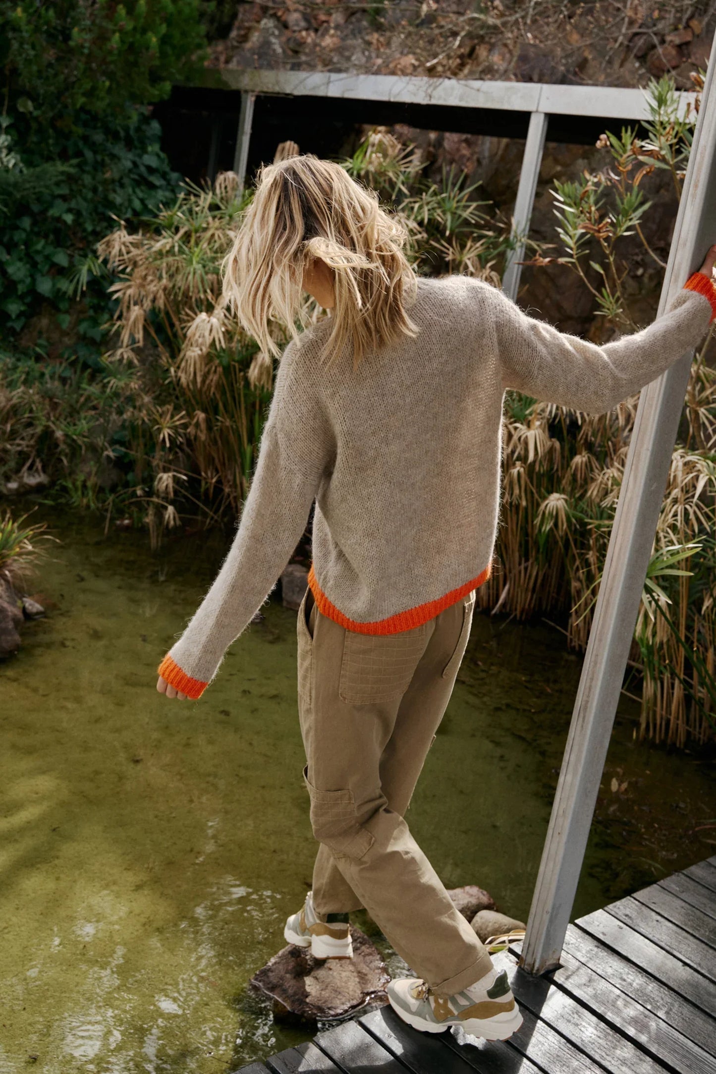 Woman in cozy beige sweater with orange trim, cargo pants, and sneakers walking over pond on wooden bridge