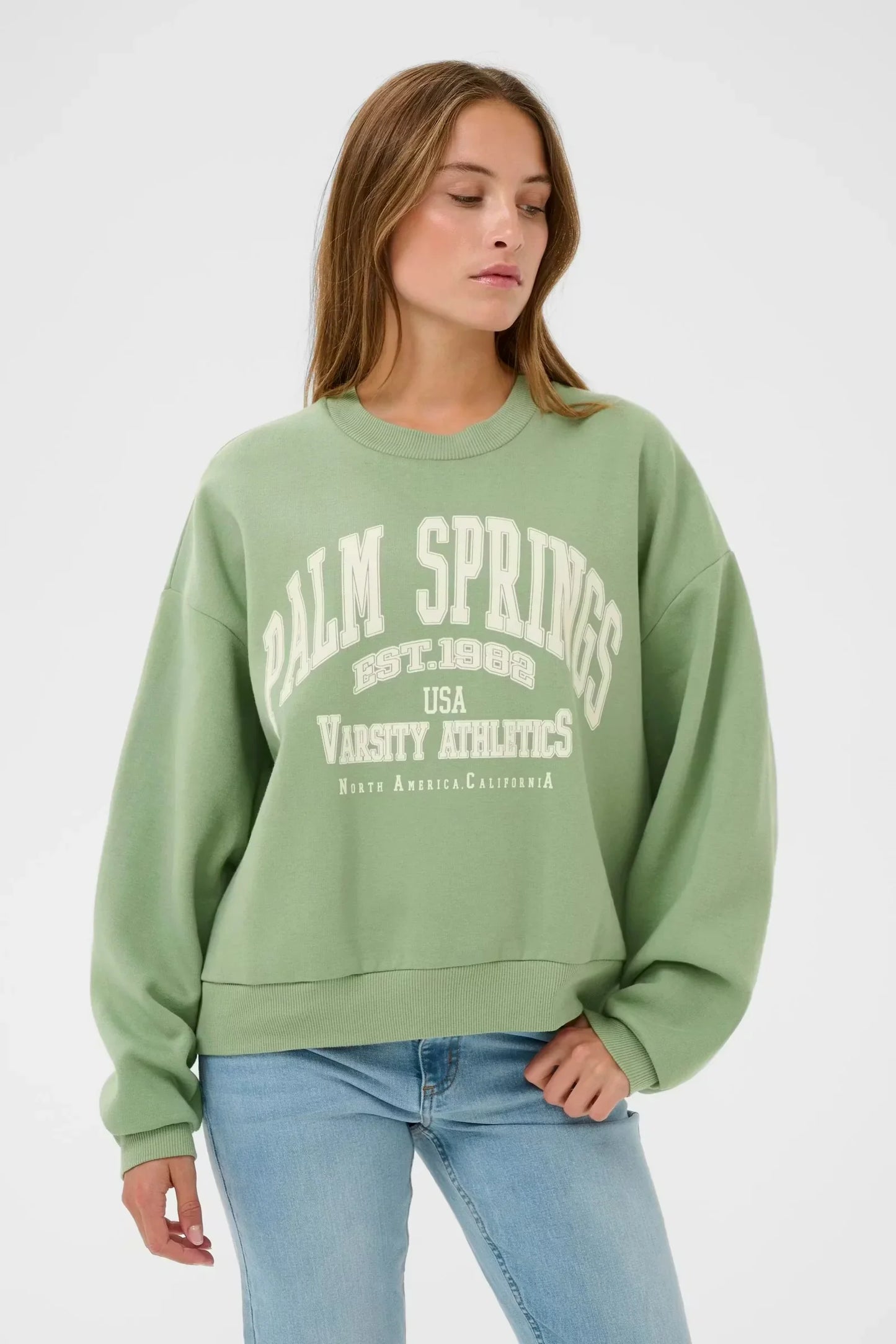 Woman wearing green Palm Springs varsity graphic sweatshirt with light wash jeans, casual women's fashion