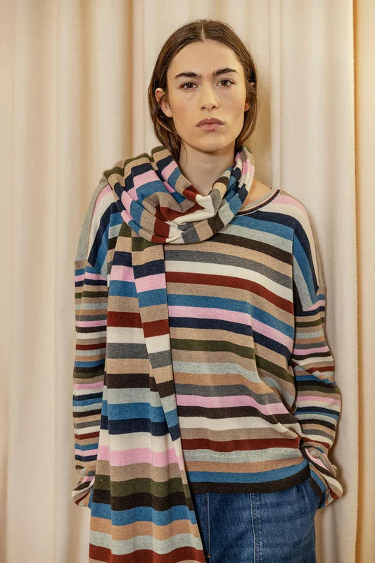 Woman wearing colorful striped knit sweater and scarf, standing indoors against beige curtains