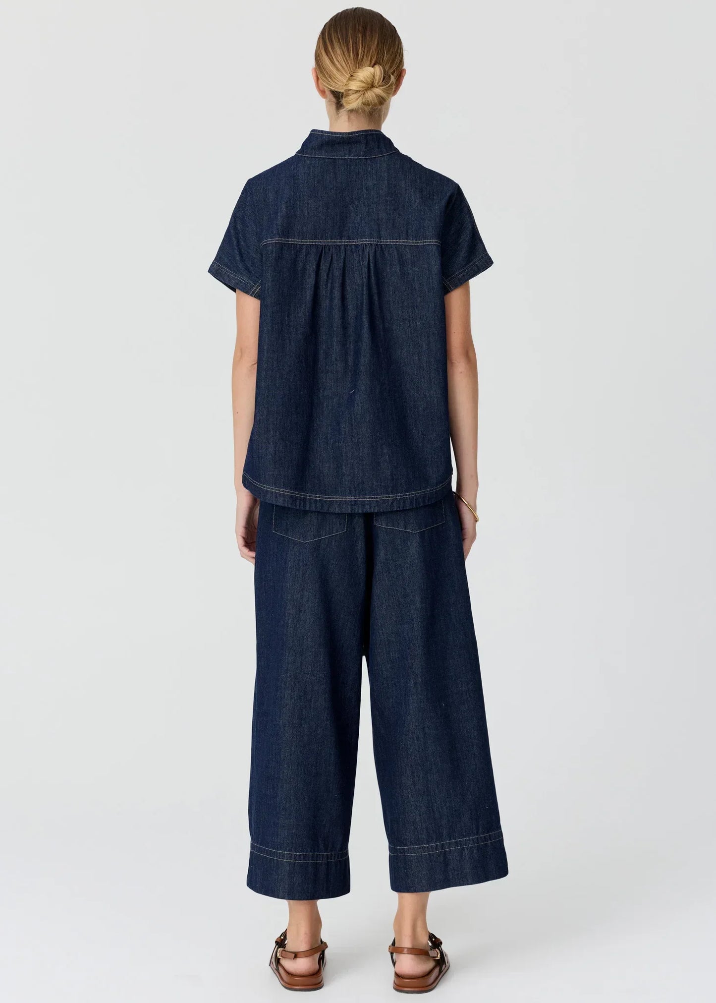 Back view of a woman wearing a dark wash denim short-sleeved shirt and matching wide-leg pants.