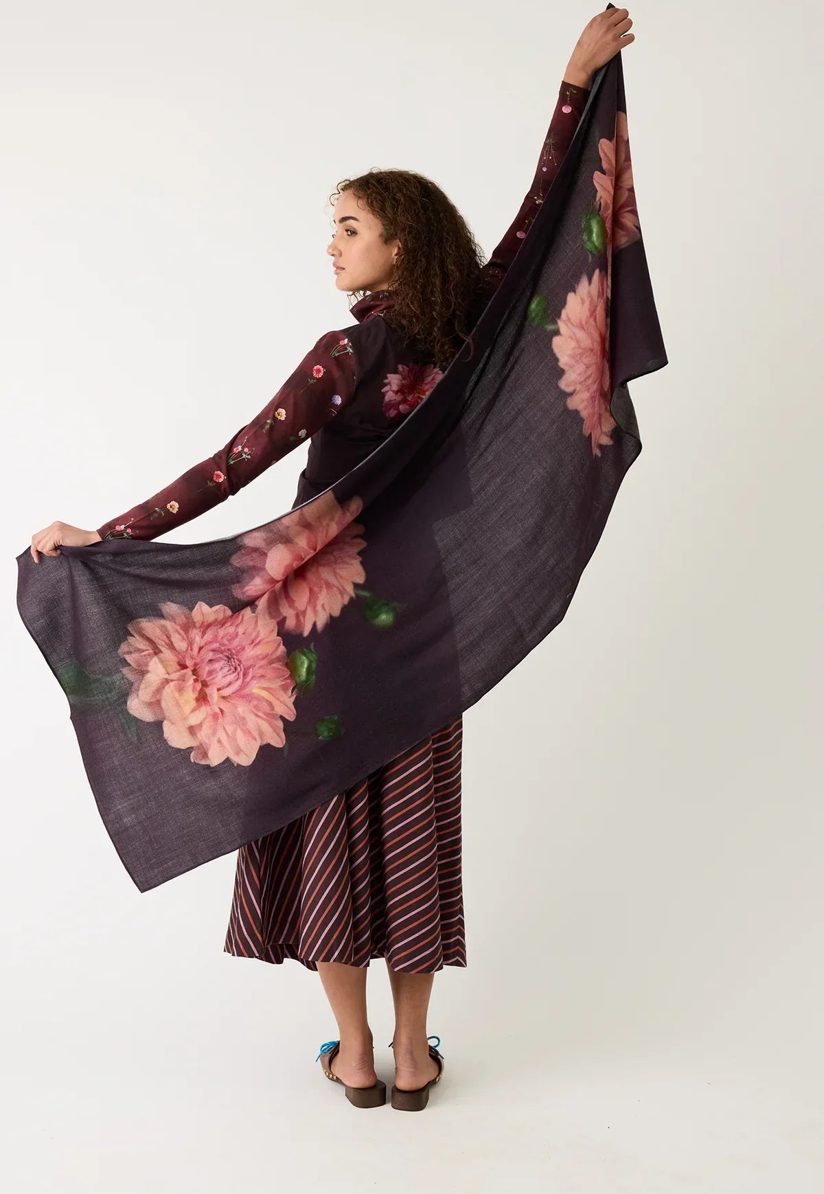 Woman wearing floral print scarf and striped skirt, holding scarf wide against white background