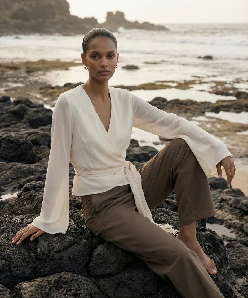 Woman in cream wrap blouse and brown trousers sitting on volcanic rocks by the ocean.