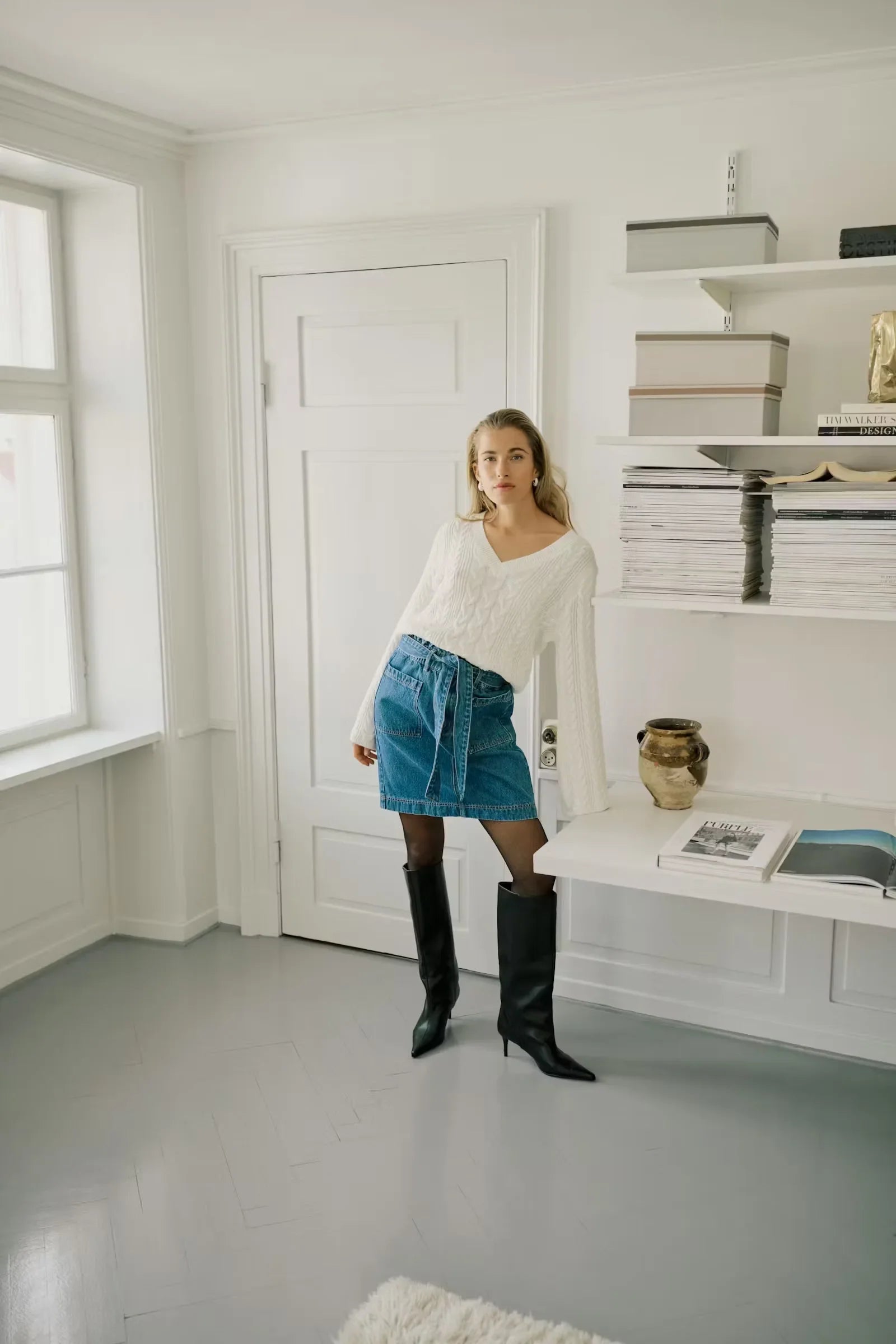 Woman in white cable knit sweater, blue denim mini skirt, and black knee-high boots in bright room