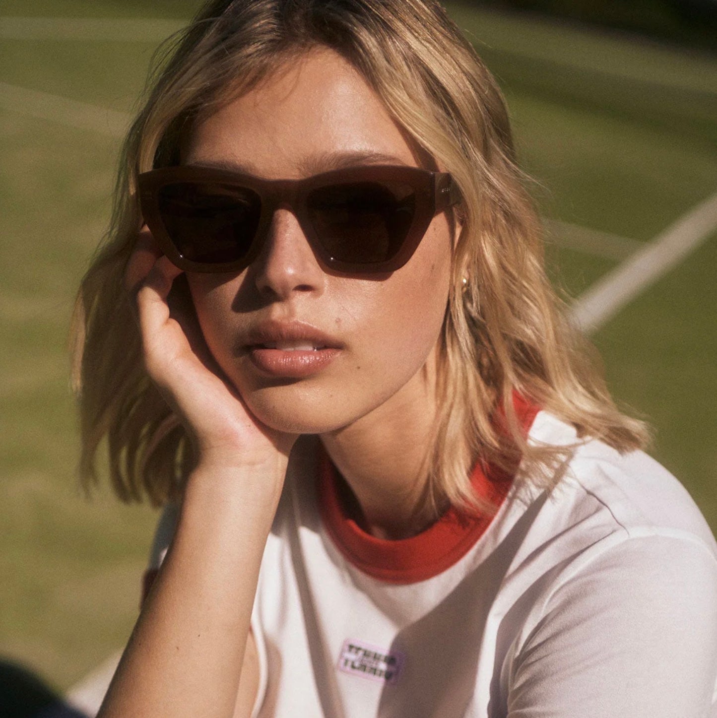 Woman wearing stylish brown sunglasses and white tee with red collar, posing outdoors on grass