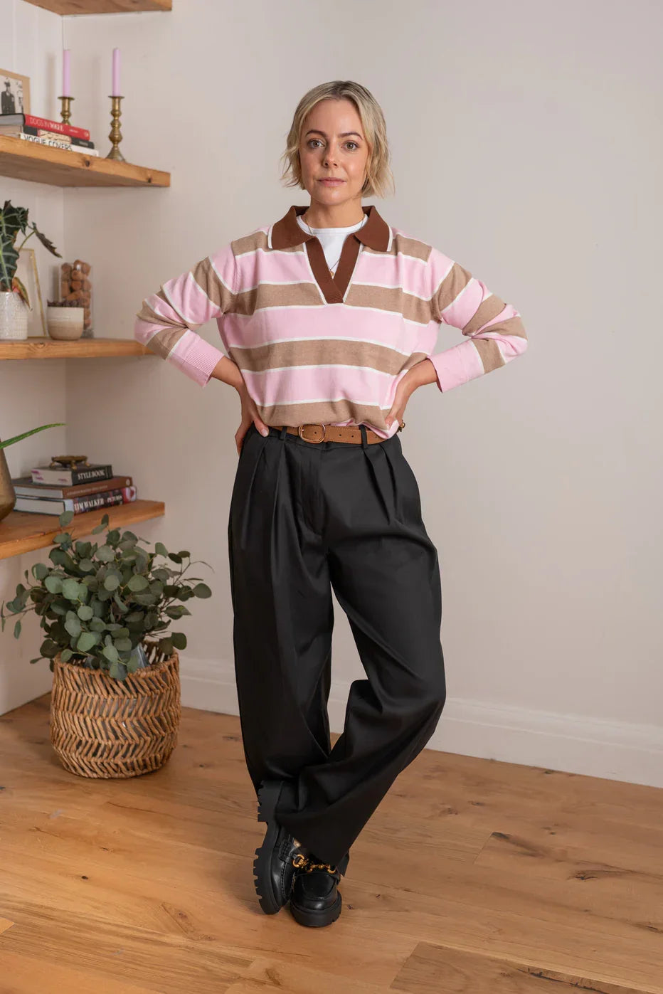 Woman wearing a pink and taupe striped rugby top with black pleated trousers and loafers indoors