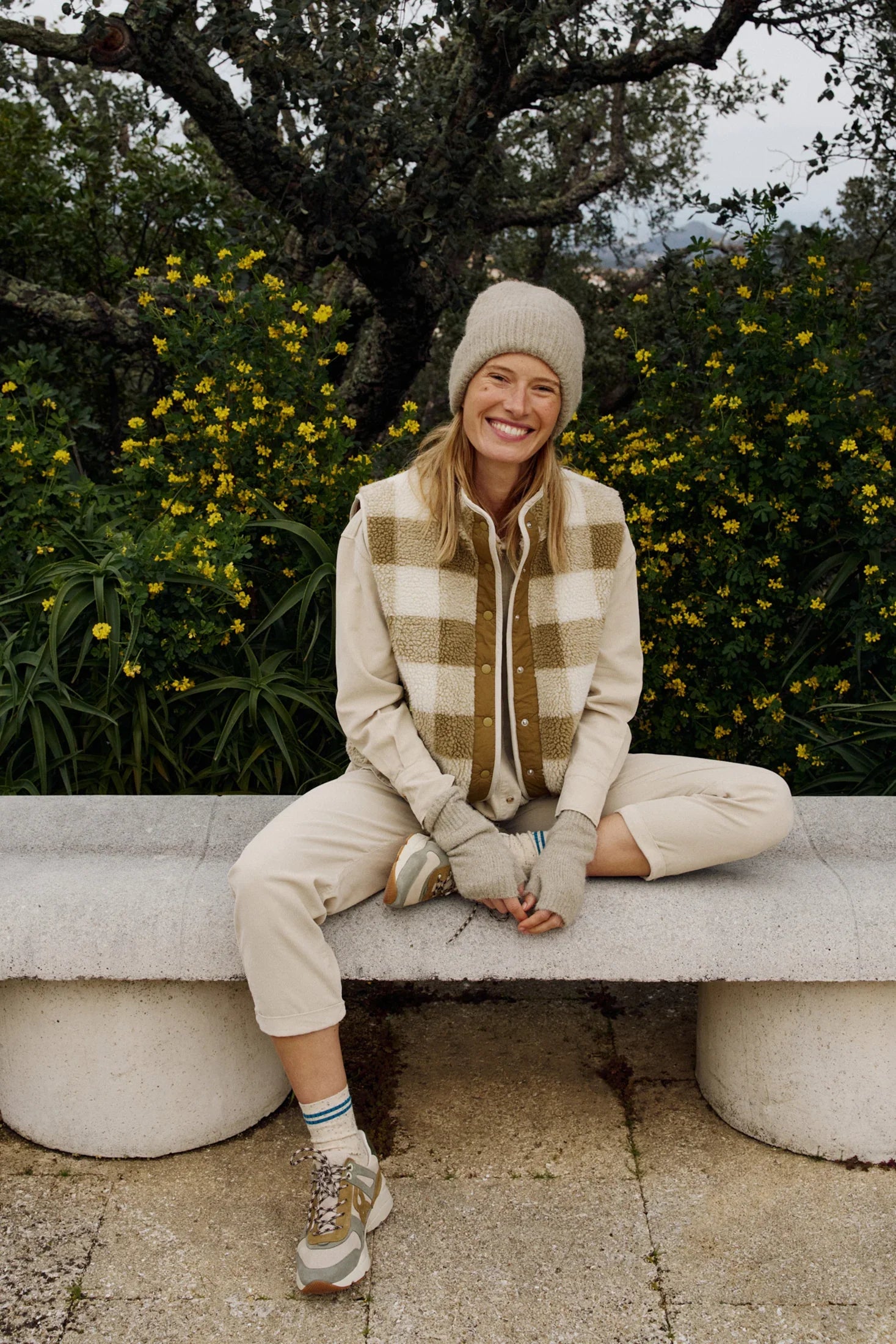 Woman in cozy plaid vest, beanie, and sneakers sitting on bench outdoors with yellow flowers