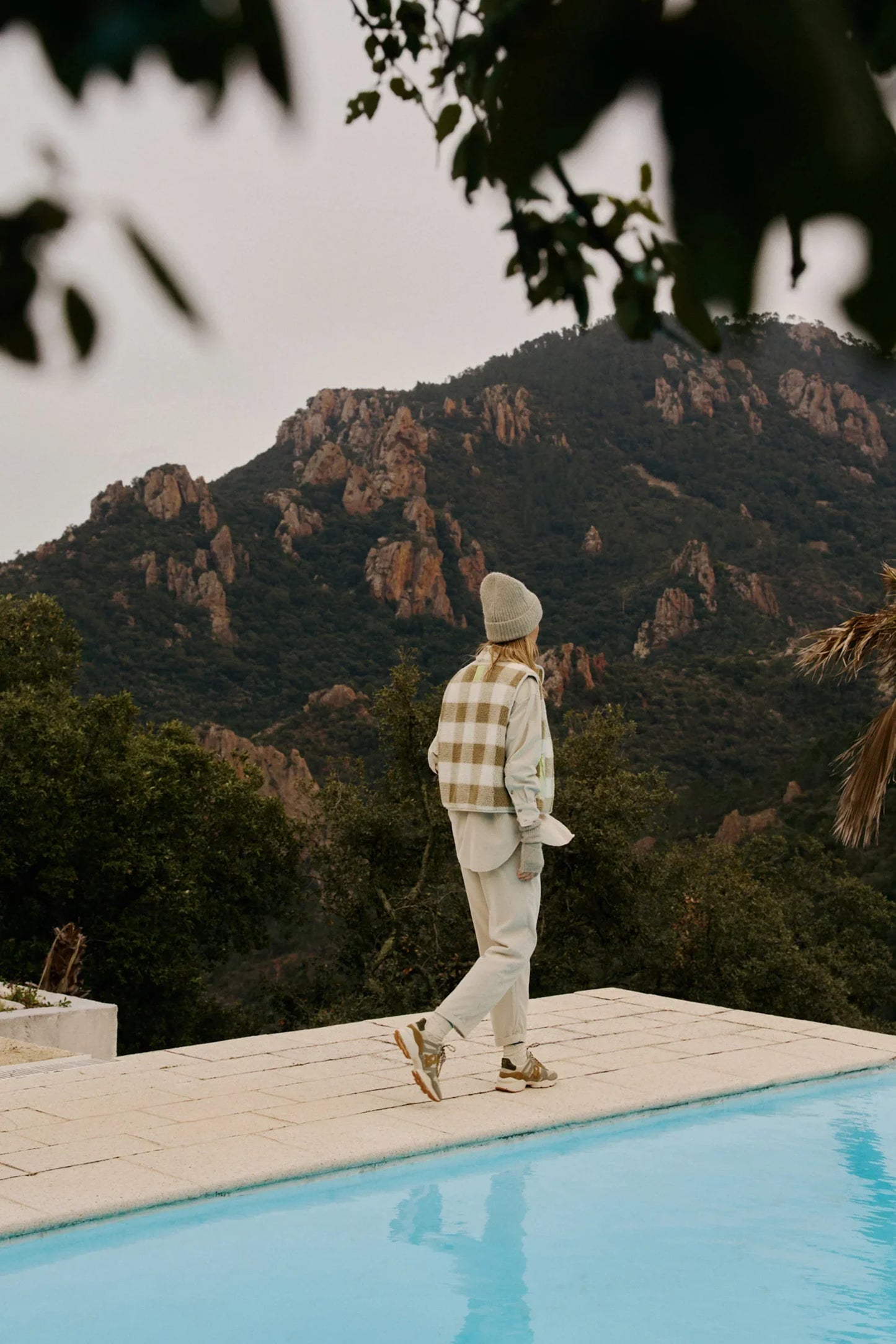 Woman in plaid vest and beanie walking by pool with mountain view, stylish women's fashion