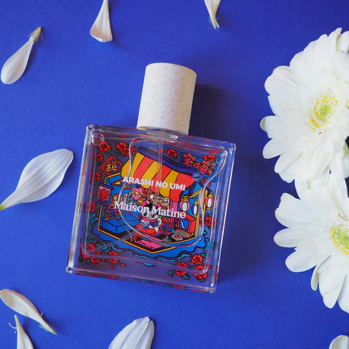 Maison Matine Arashi No Umi perfume bottle with vibrant label, white flower petals on blue background