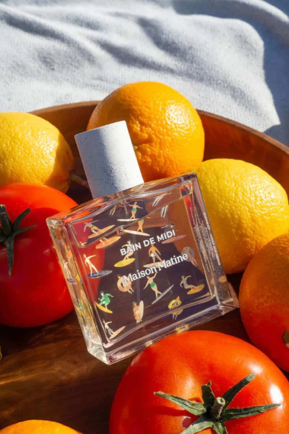 Maison Matine perfume bottle with surfers, surrounded by oranges, lemons, and tomatoes