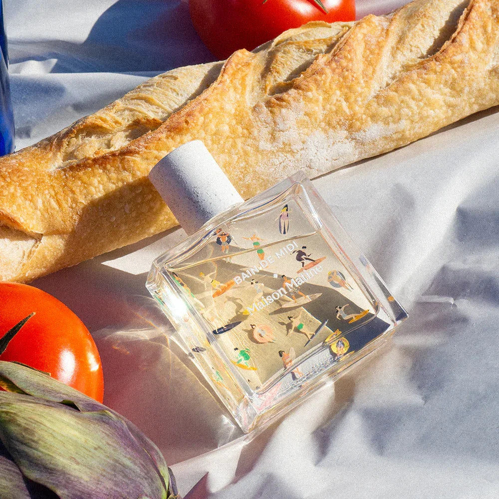 Clear perfume bottle with beach-themed design beside fresh baguette and tomatoes