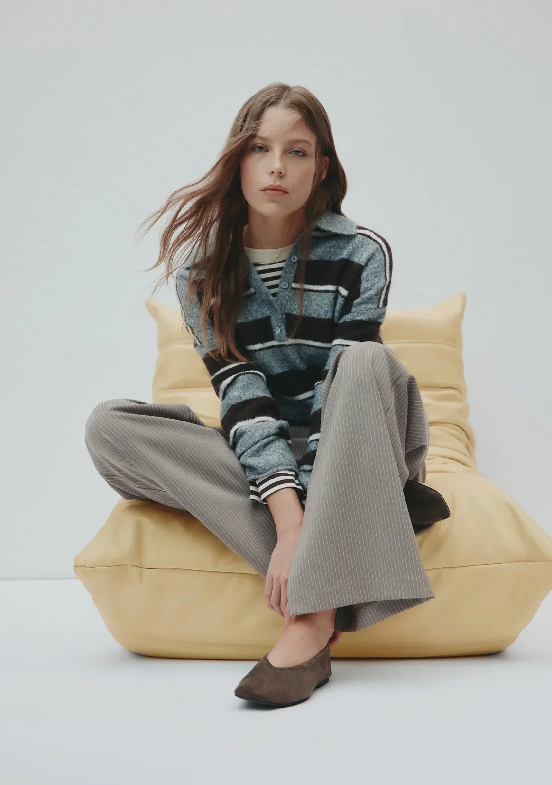 Woman in striped cardigan, pinstripe pants, and suede flats sitting on yellow bean bag
