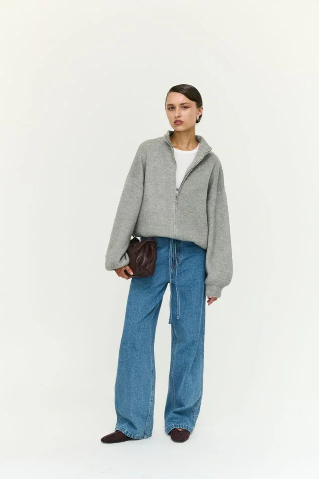Woman in grey zip-up sweater, wide-leg jeans, and brown loafers, holding leather clutch